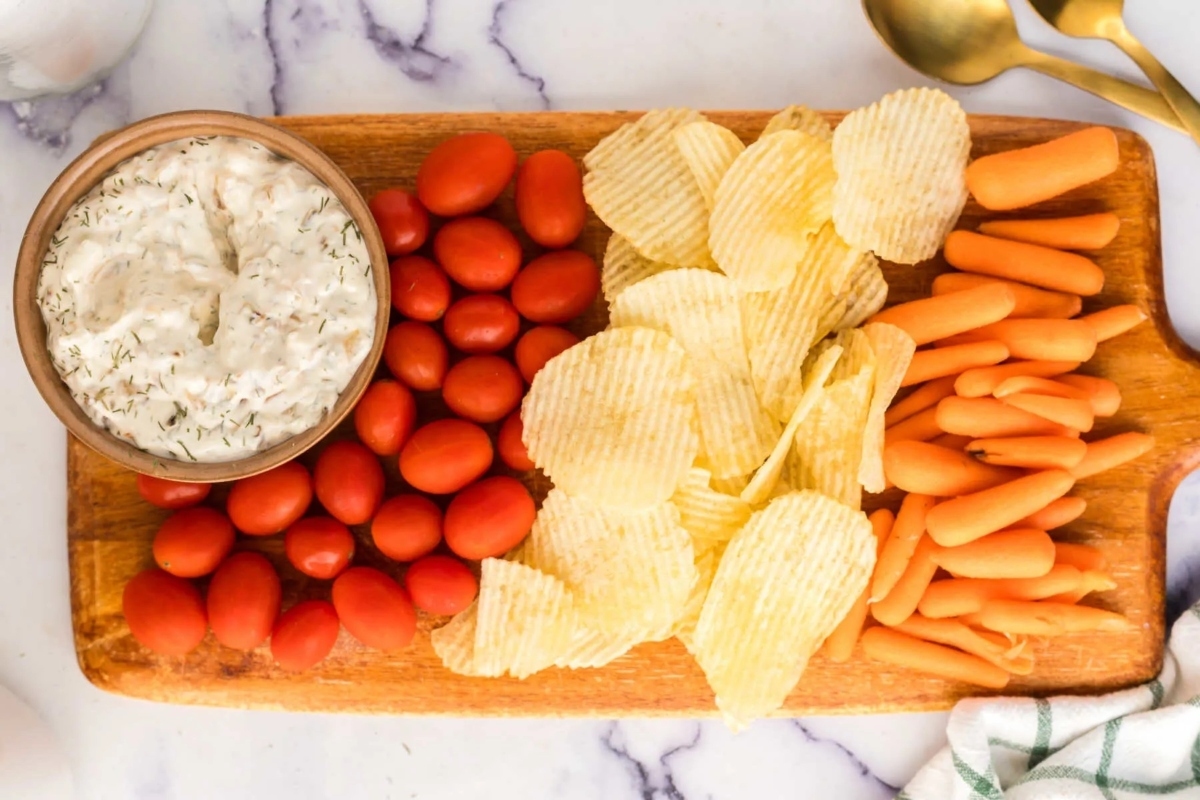 A wooden cutting board with carrots and chips served with a creamy dip.