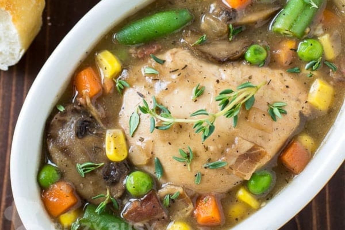 Slow cooker chicken stew with vegetables and bread on a wooden table.
