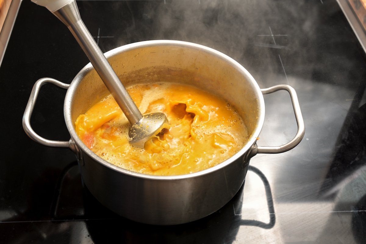 Vegetarian vegetable soup from Hokkaido or red kuri squash is pureed with an immersion blender in a steel pot on a black stove, cooking in autumn, copy space, selected focus, narrow depth of field