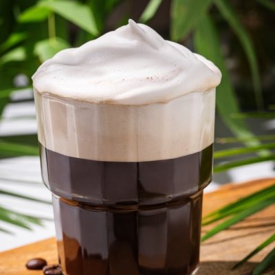 Iced coffee in a glass with foam on a wooden board on a light background with coffee beans and morning shadows in tropical branches. Summer refreshment concept. Front view.