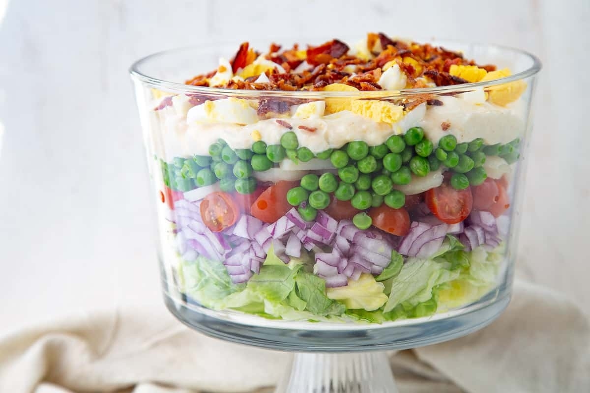 A layered salad with lettuce, red onion, tomatoes, peas, cheese, eggs, and bacon, presented in a clear glass bowl.