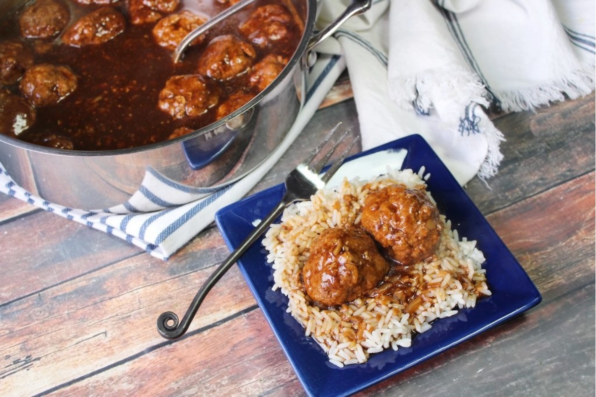 A plate of rice topped with meatballs in sauce, with a pot of extra sauce and meatballs in the background.
