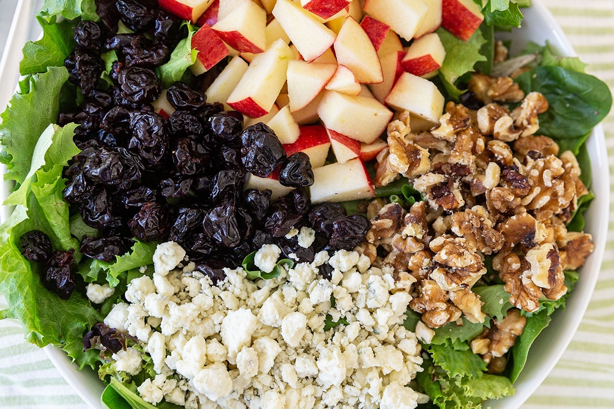 Fresh salad with leafy greens, topped with apple slices, raisins, walnuts, and crumbled cheese.