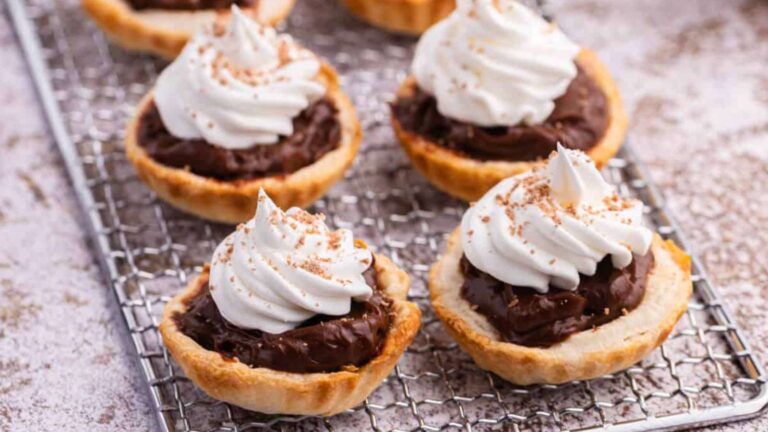 Chocolate tarts topped with whipped cream and a dusting of cocoa on a wire rack.