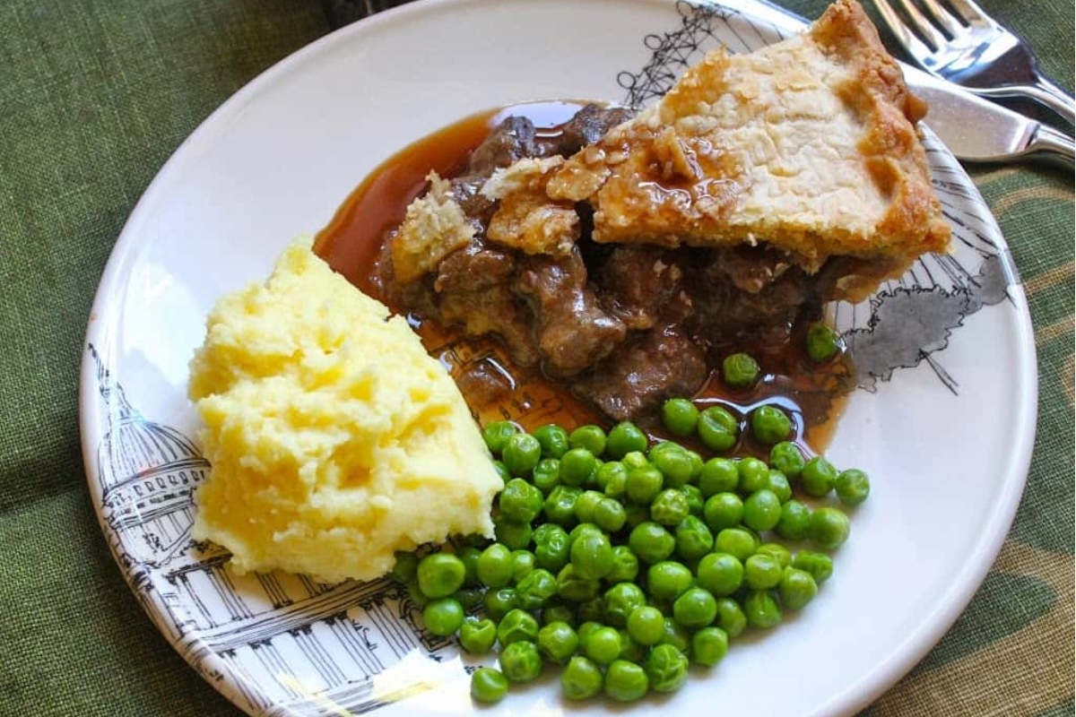 A plate of food consisting of mashed potatoes, green peas, a piece of pie, and meat with gravy.