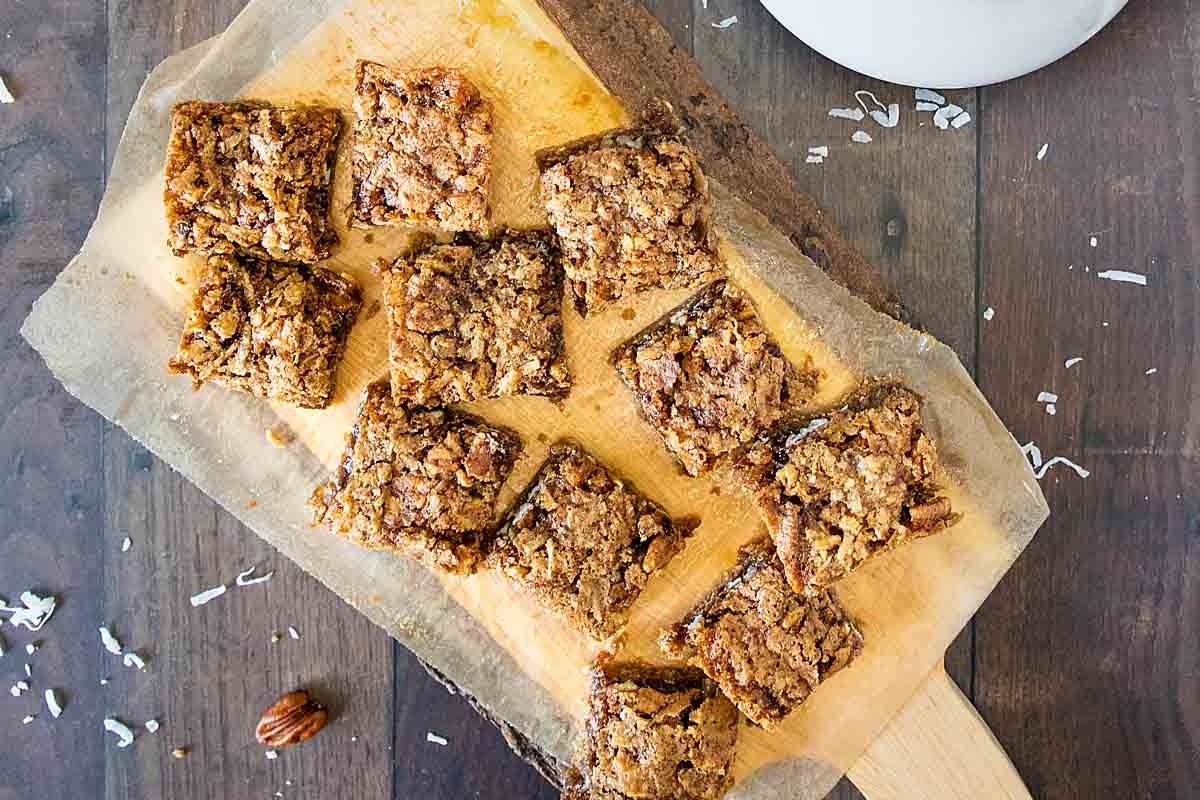 Freshly baked pecan bars on parchment paper with scattered nuts and coconut flakes on a wooden surface.