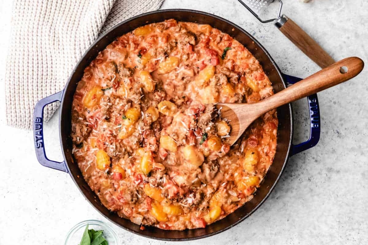 A stovetop casserole with tomatoes, cheese, and meat in a blue cast iron skillet with a wooden spoon.