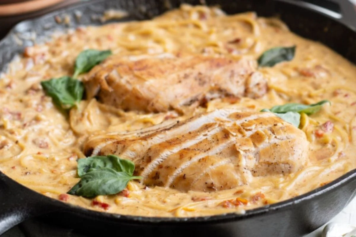 Creamy pasta with two grilled chicken breasts and basil leaves in a cast iron skillet.