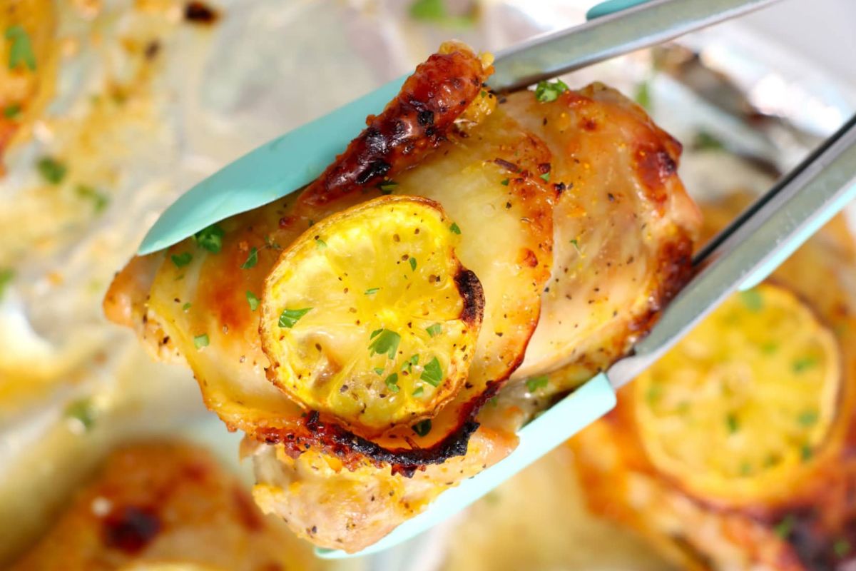 23 Finger-Lickin' Good Chicken Thigh Recipes That Are Simply Irresistible 10 Close-up of metal tongs holding a piece of roasted chicken topped with a slice of lemon and garnished with chopped herbs, perfect for those exploring delicious lemon recipes.
