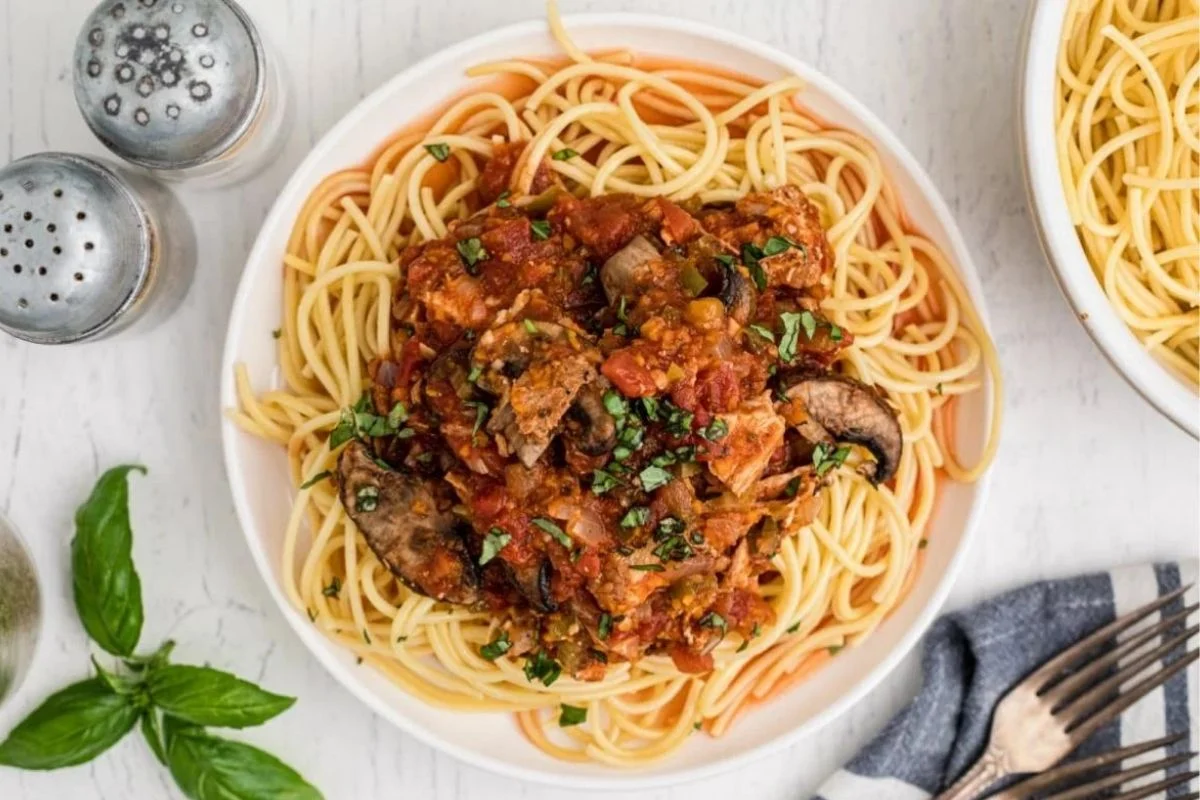 A plate of spaghetti topped with a tomato-based sauce containing mushrooms and herbs, meticulously crafted using slow cooking techniques. Salt and pepper shakers, basil leaves, and a cloth napkin are nearby.