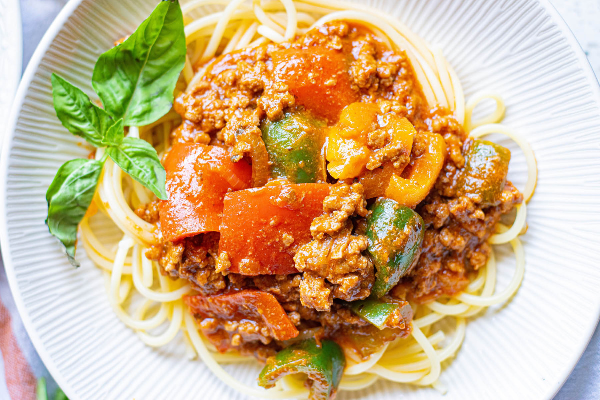 A plate of Italian cuisine featuring spaghetti noodles topped with rich meat sauce, colorful bell peppers, and garnished with fresh basil leaves.
